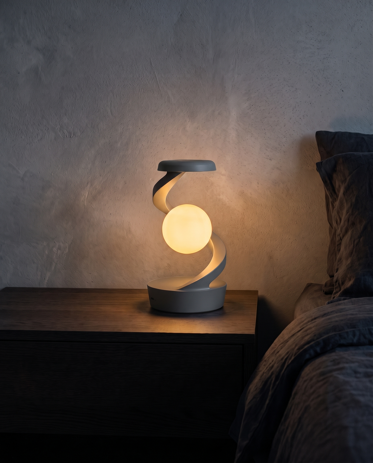 Orbit Lamp in grey, glowing warm white on a bedside table